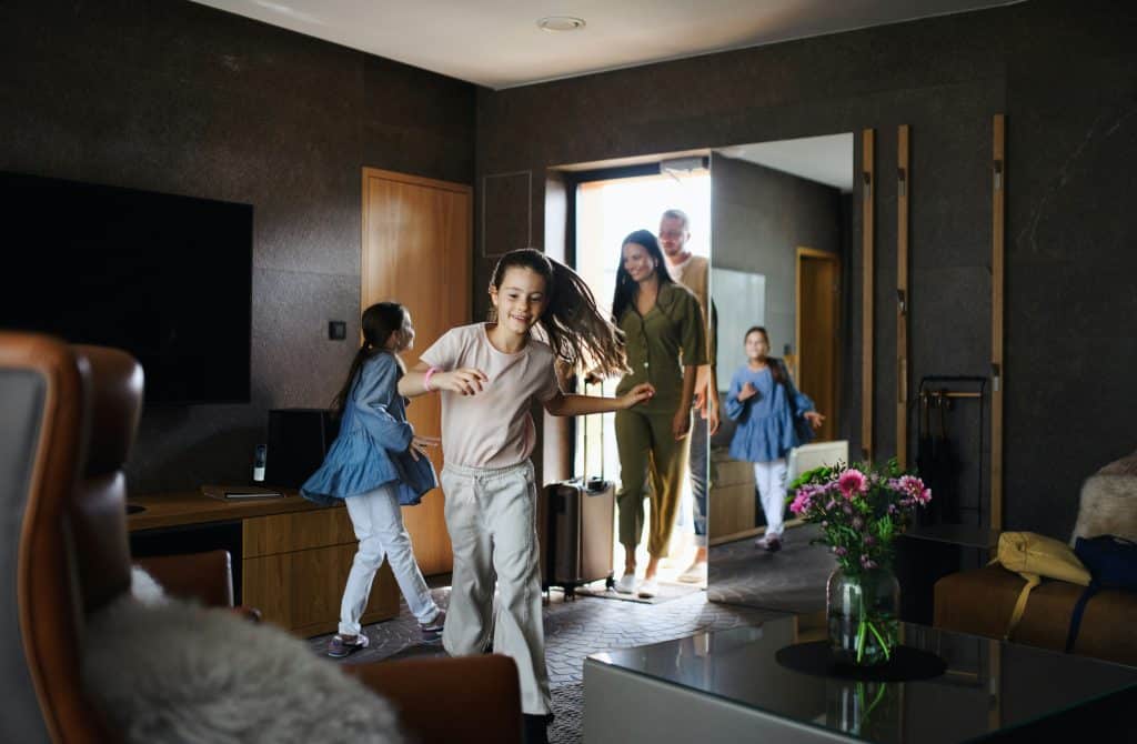 Family arriving in their hotel room with kids running excitedly ahead, capturing the joyful chaos of holiday travel and the need for reliable hotel childcare support