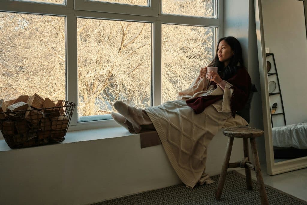 Mother sitting by a window with a warm drink, enjoying a quiet winter moment that reflects calm preparation for the season ahead