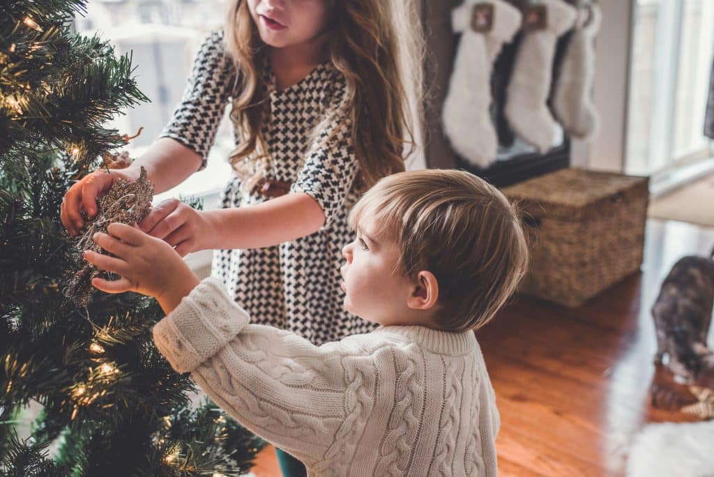 Two young children decorating a Christmas tree together in a cozy living room, capturing the warm, organized holiday moments families enjoy when childcare support helps keep Decembe