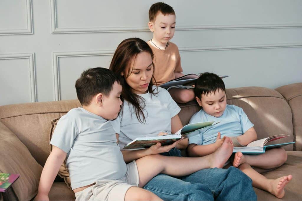 An adult sitting on a couch reading with three children, each holding a book in a calm living room