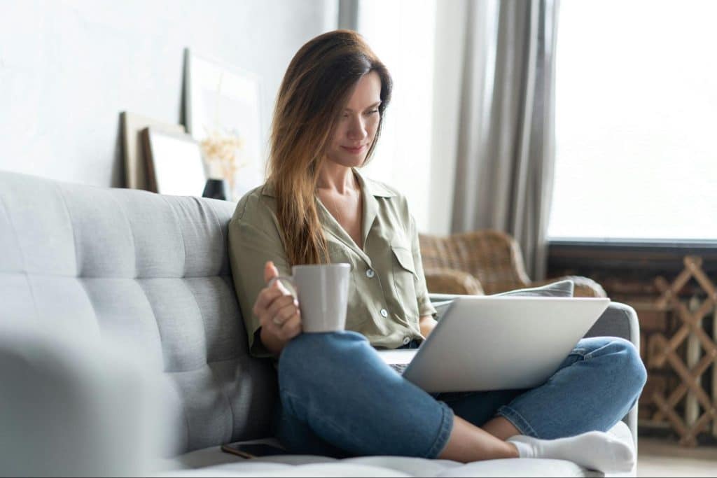 An adult sitting on a couch with a laptop and a mug, working calmly in a softly lit living room
