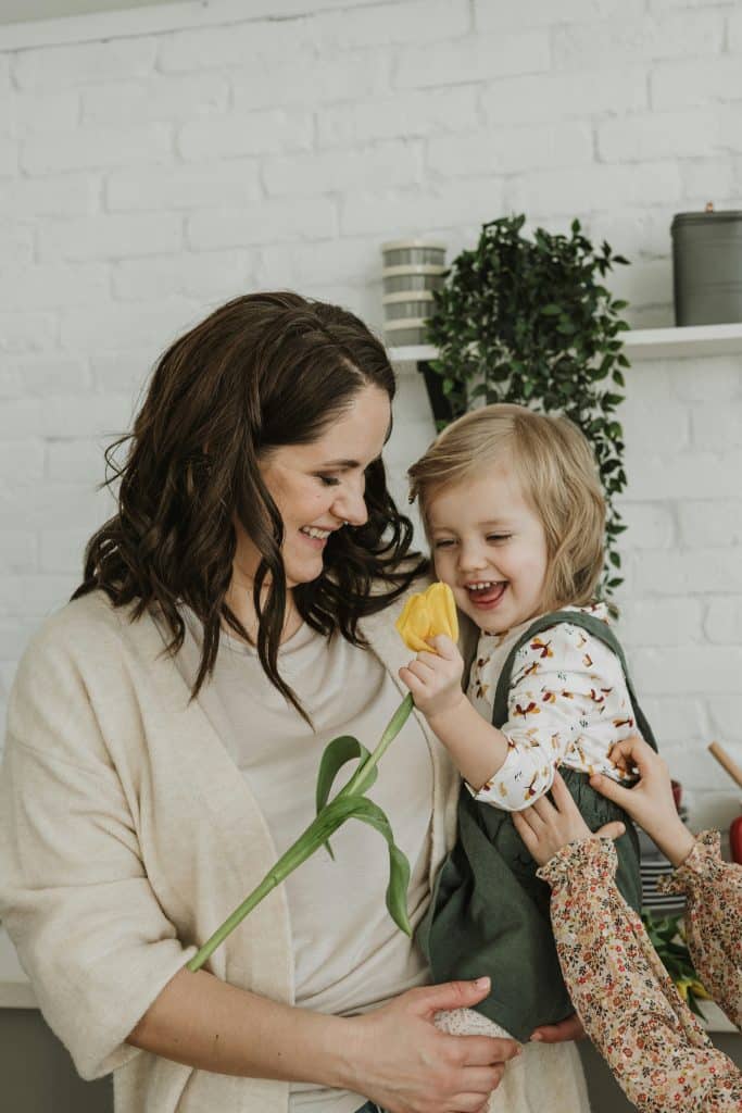 Mother holding smiling toddler with a yellow tulip in a calm home setting, illustrating steady family support during school break