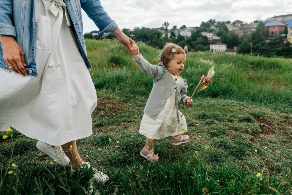 Mother holding toddler’s hand while walking through a grassy field during spring break, illustrating steady childcare support and preparedness