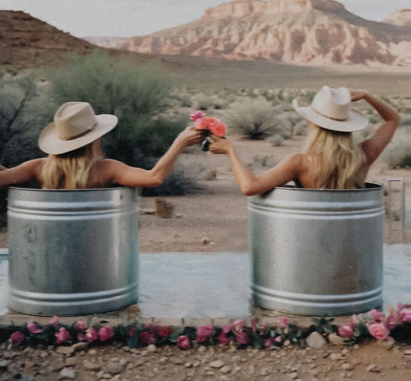 Two women sitting outdoors in metal tubs, exchanging flowers, representing connection, care, and women’s wellness