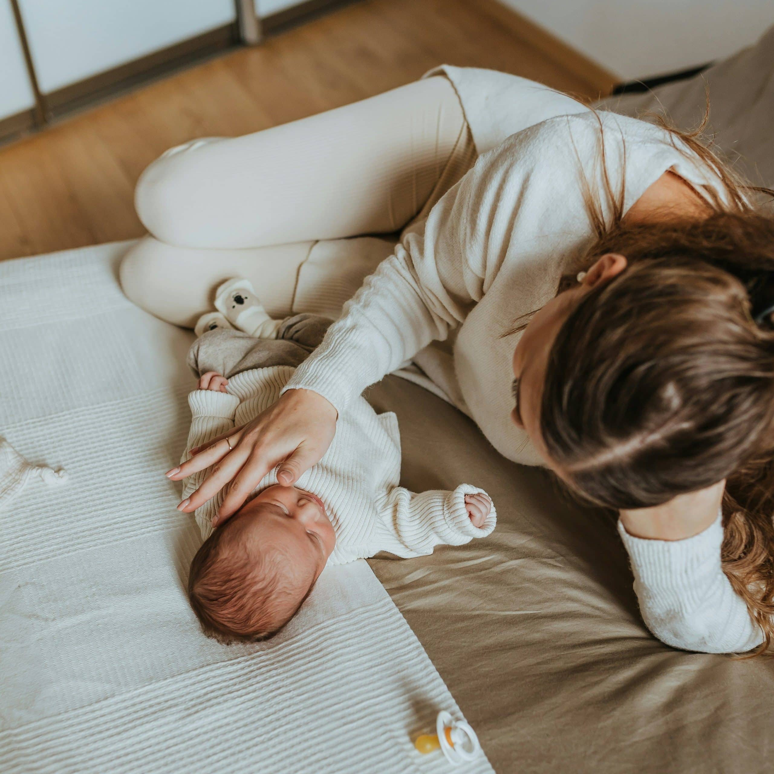 Mother lying on a bed beside her sleeping newborn, gently resting her hand on the baby's head