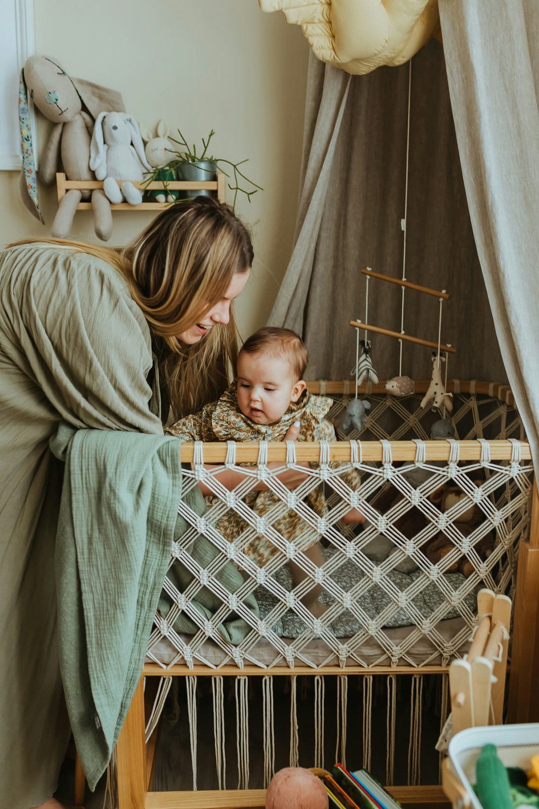 Mother smiling at her baby standing in a macrame crib in a cozy nursery
