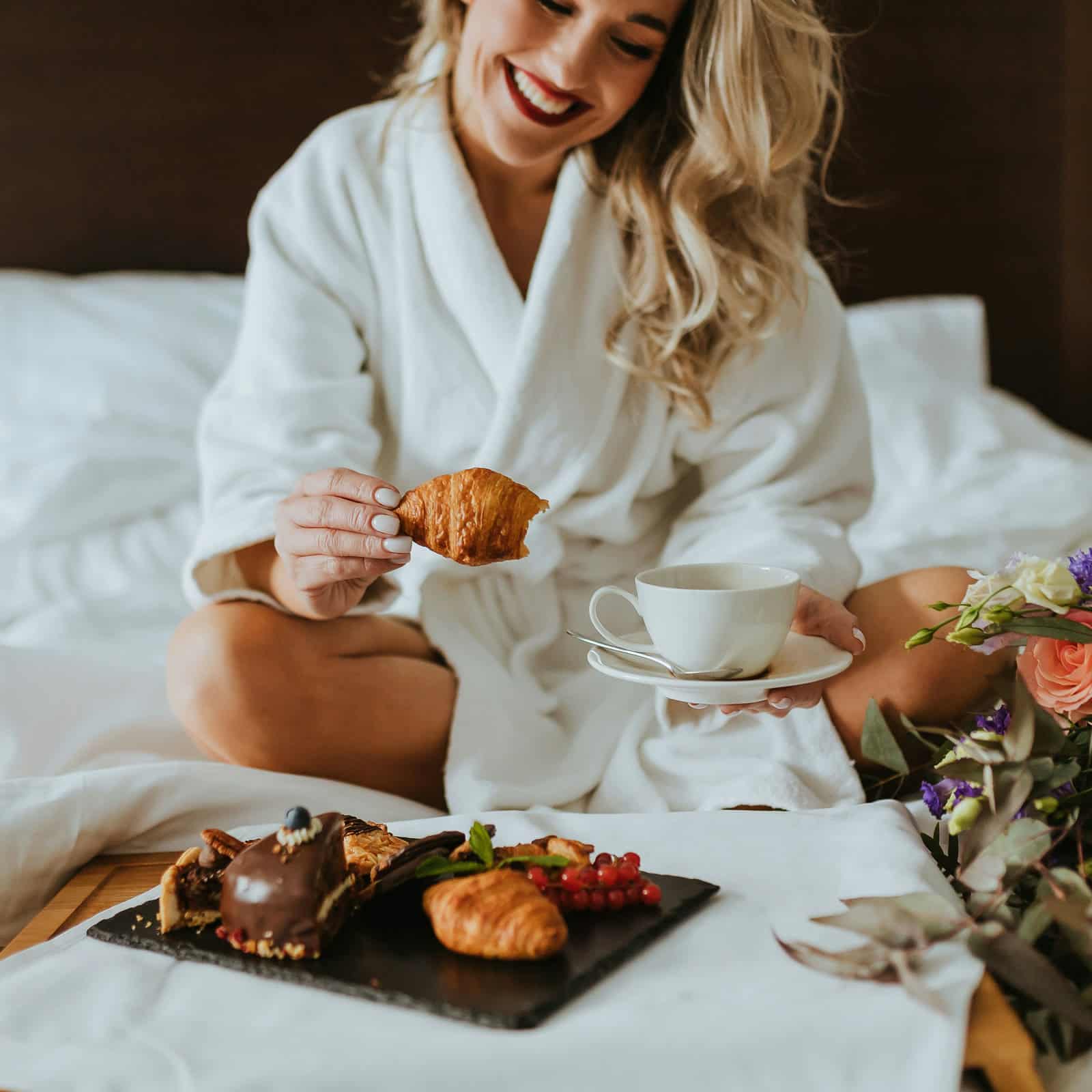 Smiling woman in a white robe enjoying breakfast in bed with coffee and pastries
